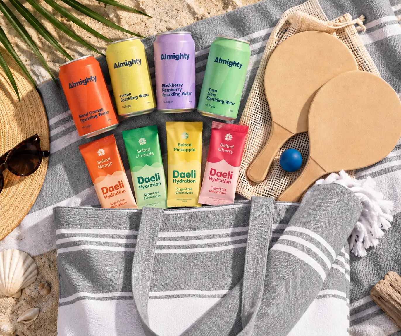 The Beach Day Gift Set flat lay: grey Hummum beach bag and hammam towel, Raquet paddle set, four Almighty Sparkling Waters and four Daeli Hydration sachets